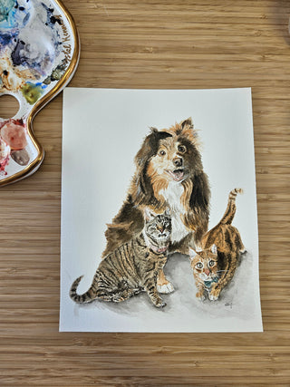 Watercolor representation of a dog and 2 cats placed on a wooden desk with a watercolor palette in the corner.