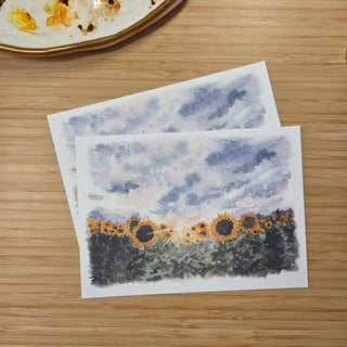 Two square art prints featuring a watercolor landscape of sunflowers under a cloudy blue sky, displayed on a light wood tabletop with a partial ceramic dish visible in the top left corner.